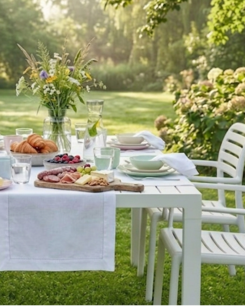 Weßer Frühstückstisch mit Blick in einen parkähnlichen Garten mit Hortensien und einer von vielen Sträuchern und Bäumen eingegrenzter großer Rasenfläche. Weiße Stühle und Tisch von Nardi,  weiße Tischläufer und Stoffservietten mit Hohlsaum von Lindhorst 1881.