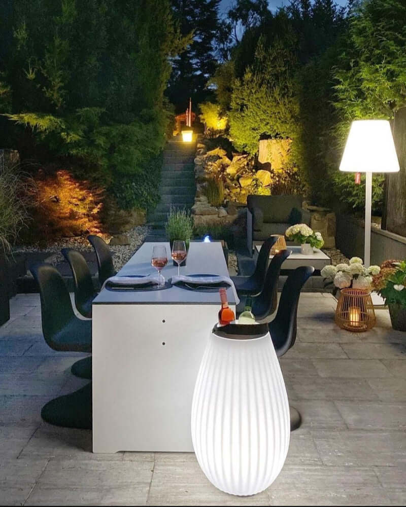 The Joouly vor Tisch im Garten mit Treppe im Hintergrund weiß leuchtend.