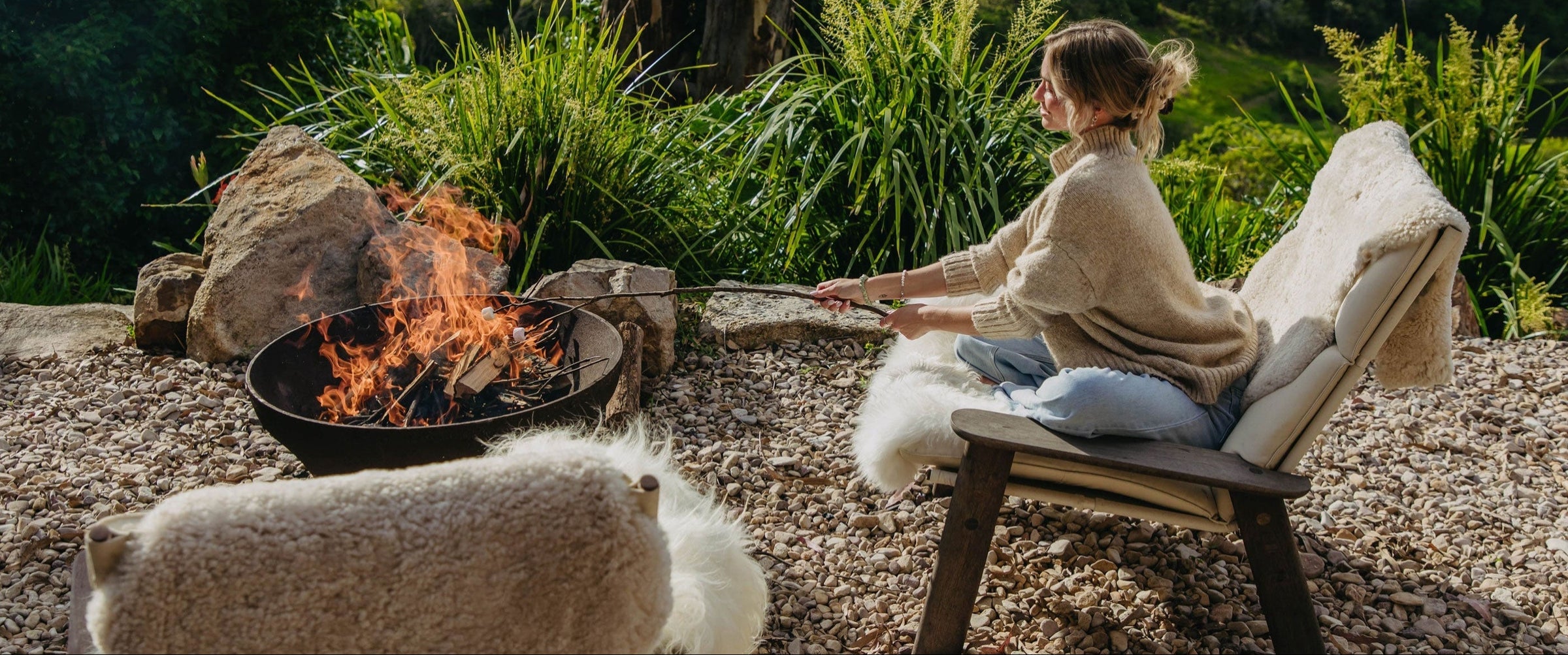 Frau sitzt im Freien am Feuer auf Lammfellen und grillt eine Wurst am Spieß.