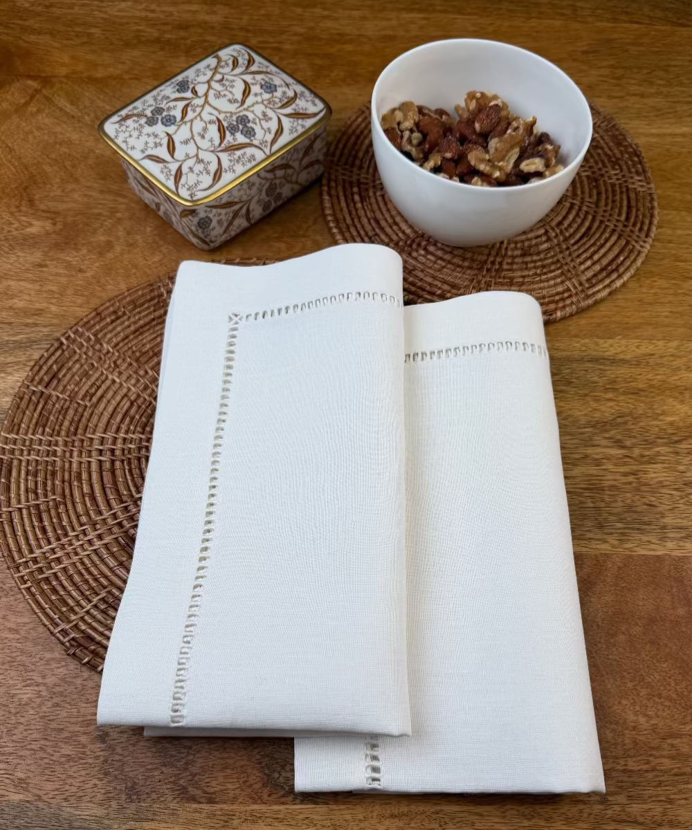 Leinenserviette Riva 50x50cm weiß mit geflochtenem Untersetzer und weißer Nussschale dekoriert.
