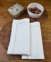 Leinenserviette Riva 50x50cm weiß mit geflochtenem Untersetzer und weißer Nussschale dekoriert.