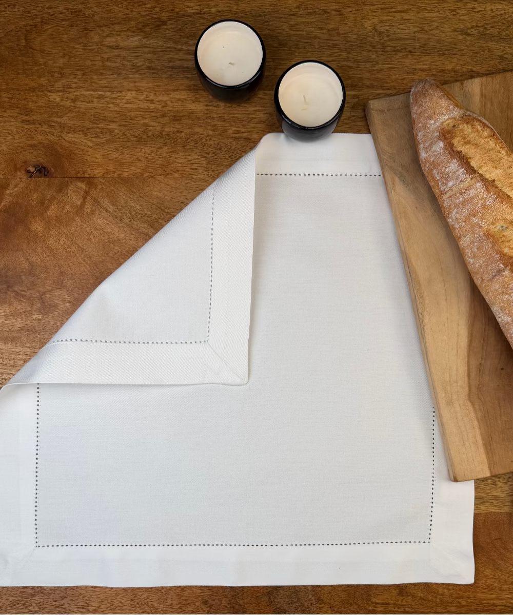 Stoffserviette Como weiss 45x45cm mit Hohlsaum und 4cm Bordüre auf Holztisch mit Holzbrett und Baguette. 