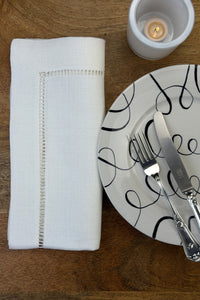 Stoffservietten Finca 50x50cm handgestickt mit Hohlsaum auf Holztisch mit Teller, Besteck und Teelicht eingedeckt.