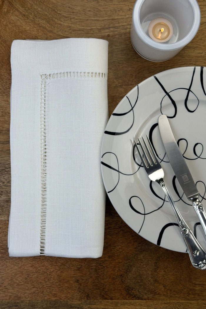 Stoffservietten Finca 50x50cm handgestickt mit Hohlsaum auf Holztisch mit Teller, Besteck und Teelicht eingedeckt.