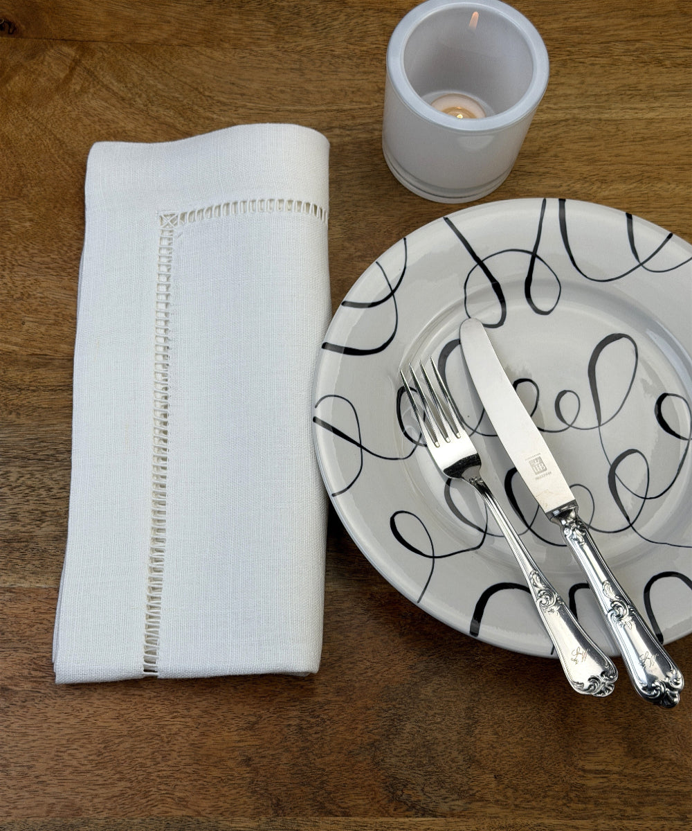 Stoffservietten Finca 50x50cm handgestickt mit Hohlsaum auf Holztisch mit Teller, Besteck und Teelicht eingedeckt.