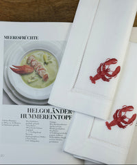 Stoffservietten Hummer 50x50cm handgestickt mit Hohlsaum auf Holztisch mit Kochbuch in Szene gesetzt.