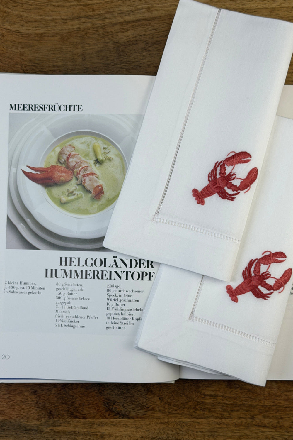 Stoffservietten Hummer 50x50cm handgestickt mit Hohlsaum auf Holztisch im Hintergrund ein Kochbuch.
