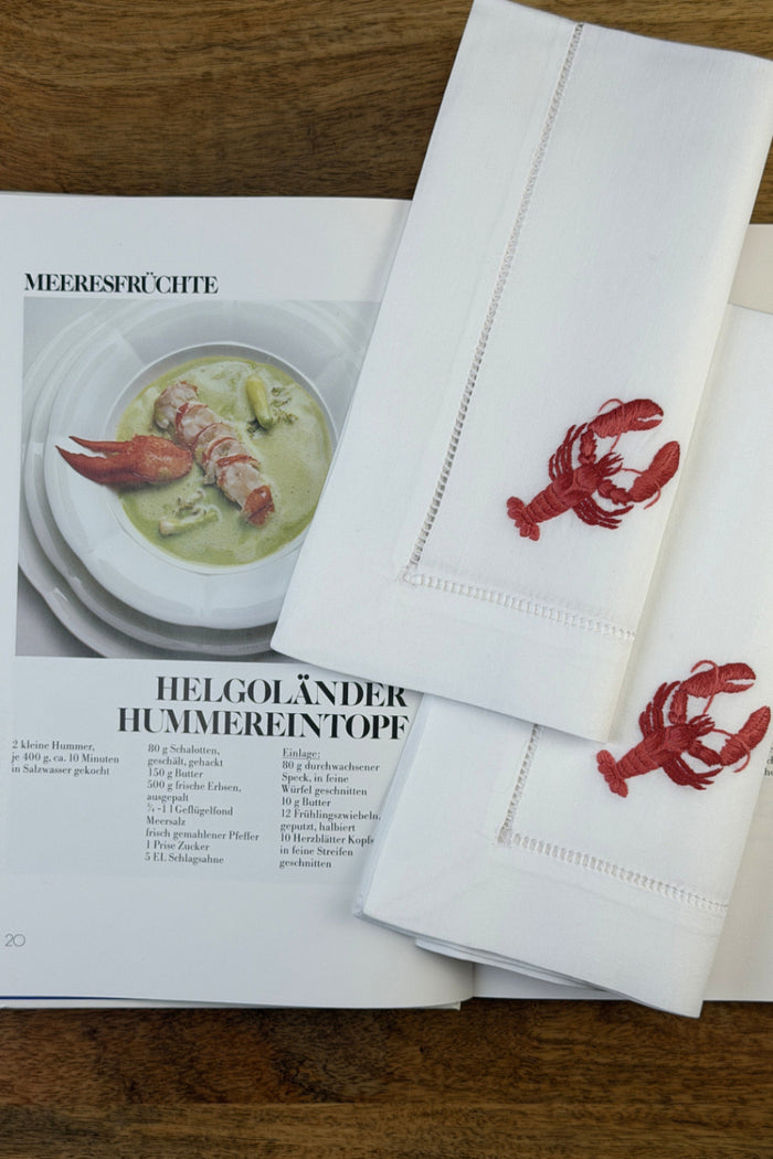 Stoffservietten Hummer 50x50cm handgestickt mit Hohlsaum auf Holztisch im Hintergrund ein Kochbuch.
