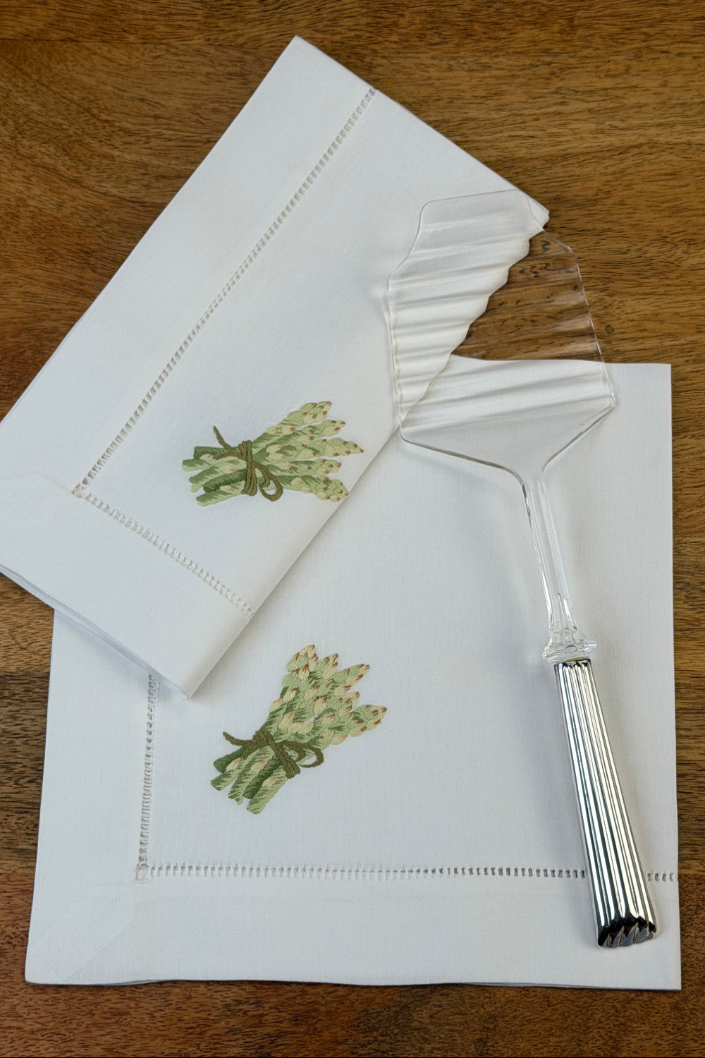 Stoffservietten Spargel 50x50cm handgestickt mit Hohlsaum auf einem Holztisch und mit einem Spargelheber dekoriert.