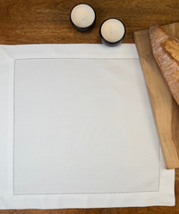 Stoffserviette Sylt weiß 45x45cm mit Hohlsaum und 4cm Bordüre auf Holztisch mit Holzbrett und Baguette.