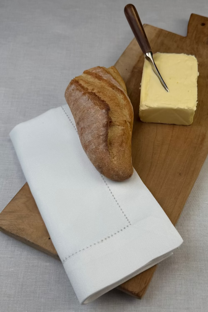 Stoffserviette Sylt 50x50cm Hohlsaum mit 4cm Bordüre auf Holzbrett mit Baguette und Butter.